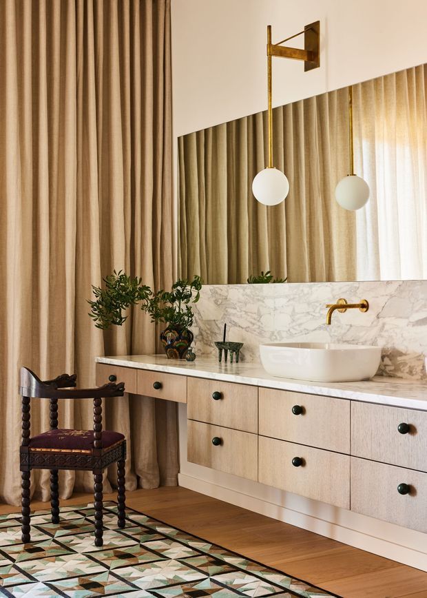 A refined bathroom vanity area with a floating light-wood drawer cabinet, white marble benchtop and integrated backsplash, vessel basin, and brushed brass wall-mounted tap. A large angled mirror reflects full-height beige curtains and globe wall lights. The space includes a patterned geometric rug, dark carved wooden chair, decorative plants, and warm timber flooring, creating an eclectic yet luxurious feel.