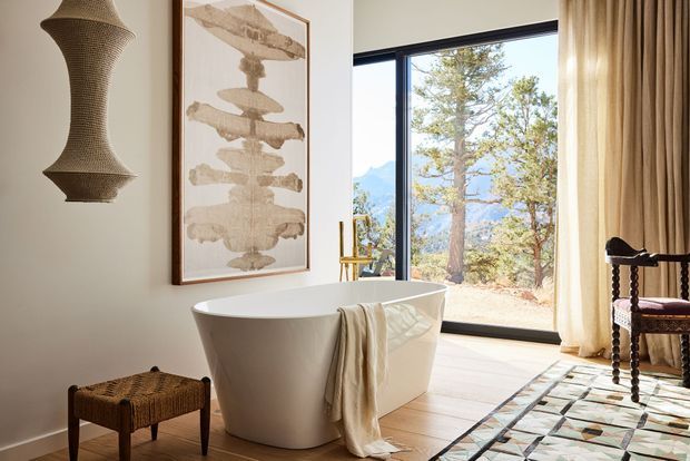 A refined freestanding bathtub occupies a bright bathroom beside expansive black-framed glazing with a mountain and tree view. The room features pale walls, light timber flooring, full-height cream curtains, a sculptural wall light, framed neutral artwork, a woven stool, and an upholstered dark wood chair. A geometric patterned rug adds a subtle graphic accent.