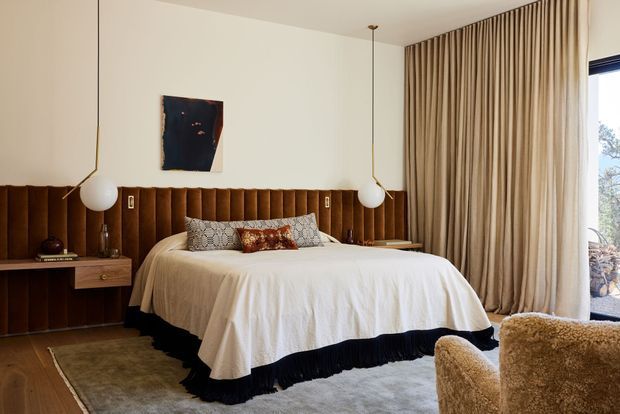 A warm, refined bedroom with a large bed dressed in white linens and a dark pleated bed skirt, set against a full-width vertical fluted timber headboard. Integrated bedside joinery and floating drawers extend along the wall, complemented by globe pendant lights, neutral curtains, a soft rug, and textured boucle seating. The room has a balanced, earthy palette with strong mid-century-inspired detailing.