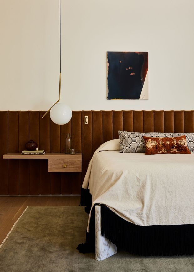 A warmly styled bedroom featuring a bed with cream linen bedding, patterned and rust-toned accent cushions, and a dramatic floor-to-ceiling channel-tufted brown upholstered wall behind the bed. A floating timber bedside table with drawer, books, glassware, and a sculptural wall-mounted globe lamp sits to the left. Abstract artwork, a muted olive-brown rug, and dark bed valance add depth to the intimate, refined space.