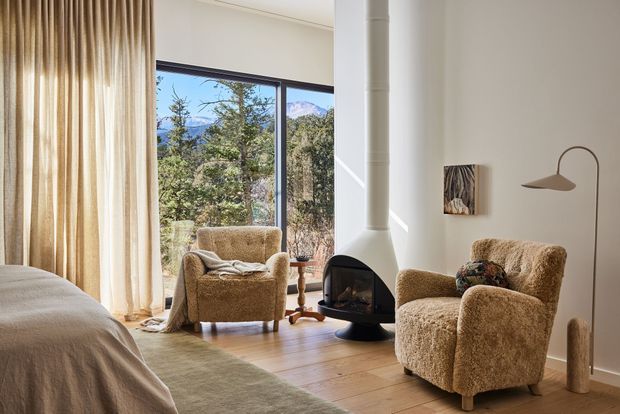 A warm, light-filled bedroom sitting area with two textured boucle armchairs arranged around a freestanding sculptural fireplace. A bed is partially visible in the foreground, with full-height curtains and large glazing opening to a wooded mountain landscape. Timber flooring, neutral furnishings, and a slim arched floor lamp create a calm, refined retreat.