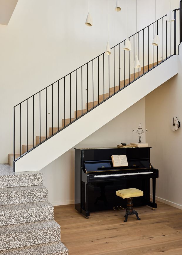 A bright, refined interior stairway with a black metal balustrade and warm timber stair treads, leading past a cluster of minimalist pendant lights. A polished black upright piano sits beneath the stairs on a timber floor, with a small upholstered piano stool and decorative wall sconce. The lower stair flight features a speckled terrazzo-like finish, while the walls and stair stringer are warm off-white.