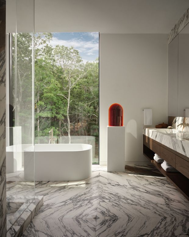 Luxurious contemporary bathroom with a freestanding white bathtub beside full-height glazing overlooking trees. The room features dramatic bookmatched marble flooring and wall surfaces, a floating dark timber vanity with a marble benchtop, integrated mirror cabinetry, brushed metal tapware, and a sculptural red-orange glass object on a pedestal.
