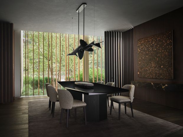 A moody, sophisticated dining room with a dark rectangular dining table, upholstered dining chairs, and a sculptural pendant light. Floor-to-ceiling glazing with vertical timber screening opens to a leafy garden, while dark built-in cabinetry, a textured feature artwork or wall panel, and dark timber flooring create a refined, intimate atmosphere.