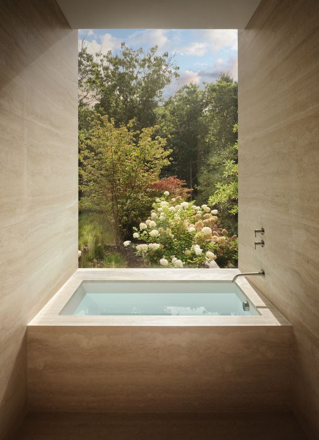 A serene, minimalist bathroom centred on a built-in soaking tub positioned in front of a large floor-to-ceiling picture window overlooking a lush garden. The walls and tub surround are clad in warm beige natural stone with subtle horizontal veining, complemented by simple wall-mounted chrome fixtures. Soft natural light and the garden view create a calm, spa-like atmosphere.