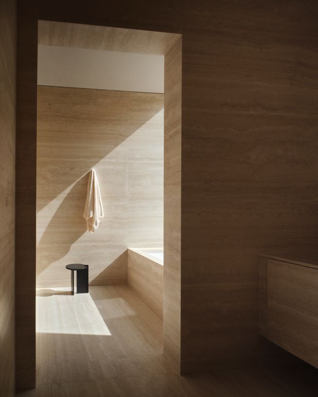 A minimalist, warm-toned bathroom clad extensively in beige natural stone, viewed through a dark timber-framed opening. The space includes a built-in stone bathtub or ledge, a small black side table or stool, and a hanging towel. Strong sunlight creates geometric shadows across the stone surfaces, while dark timber cabinetry or wall panelling occupies the foreground.
