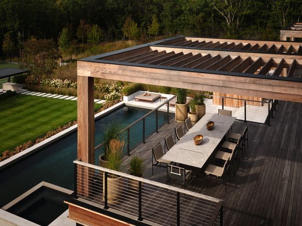 Elevated view of a contemporary outdoor entertaining deck with a long dining table beneath a timber-framed pergola featuring a translucent or open slatted roof. The deck overlooks a swimming pool and landscaped garden, with integrated railings, timber decking, potted plants, and a separate lower water feature or spa visible in the foreground. The setting is surrounded by mature woodland.