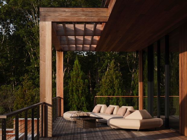 A timber-clad outdoor terrace or deck beside a modern house, surrounded by dense greenery. The space features a large cream sectional lounge, a low round timber coffee table, dark metal balustrades, and a timber pergola with a slatted translucent roof. Full-height glazing and warm timber walls form the building edge.