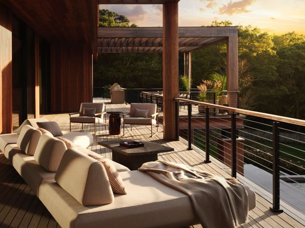A spacious elevated outdoor terrace or deck surrounded by dense greenery, furnished with a large cream sectional sofa, armchairs, a low dark coffee table and a fire bowl. Timber decking, timber posts and a pergola frame the space, while dark horizontal metal or cable-style balustrading encloses the edge. Warm late-afternoon sunlight creates a refined, relaxed outdoor entertaining atmosphere.