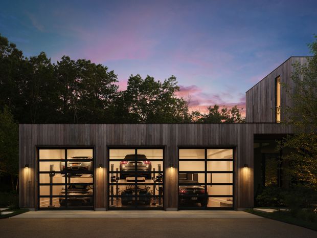 Twilight exterior view of a contemporary multi-bay garage and attached upper-level structure, clad in dark vertical timber boards. Three illuminated glazed garage doors reveal parked vehicles and storage inside, while warm wall lights contrast with the cool dusk sky and surrounding trees.