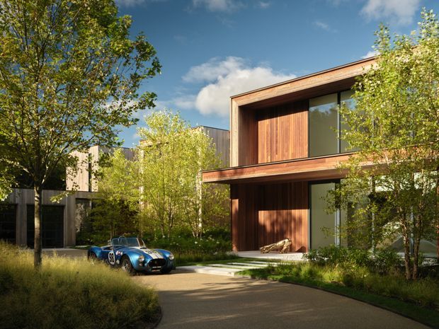 A contemporary two-storey residential building set within a landscaped suburban garden. The facade combines warm vertical timber cladding, large dark-framed glazed openings, and strong rectangular volumes with projecting roof and balcony-like edges. A curved paved driveway leads toward the entrance, with mature and young trees, ornamental grasses, and a classic blue sports car in the foreground.