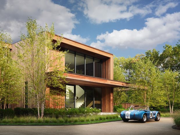Exterior view of a contemporary two-storey residence set within a leafy landscaped site. The home features warm timber cladding, strong horizontal roof planes, large floor-to-ceiling glazing, and recessed outdoor areas. A vintage-style blue sports car is parked in the foreground, while mature trees and ornamental planting surround the building.