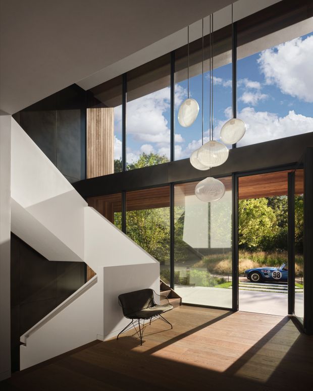 A dramatic double-height residential foyer or living area with a sculptural white staircase, dark structural beams, extensive floor-to-ceiling glazing, warm timber wall and floor finishes, and a cluster of suspended spherical pendant lights. Strong sunlight enters from the glazed facade, with trees and a vintage race car visible outside.
