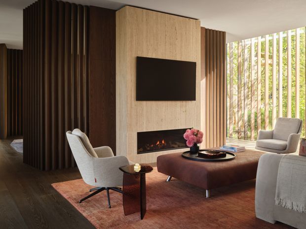 Warm, sophisticated living room with a television and linear fireplace set into a pale timber feature wall. Dark vertical timber battens define the surrounding walls and partially screen the adjacent spaces, while full-height glazed openings with vertical timber sun-screening bring in views of greenery. The room includes upholstered lounge chairs, a large deep burgundy ottoman coffee table, a sofa, side table, rug, and floral arrangement.