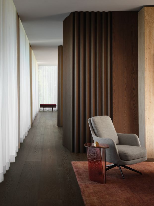 A refined interior hallway or circulation space with floor-to-ceiling sheer curtains along one side, dark timber flooring, and a dramatic vertical timber slat feature wall dividing the corridor from a small seating nook. A sculptural upholstered lounge chair and small reddish side table sit on a warm rust-toned rug. Soft natural light creates a calm, sophisticated atmosphere.