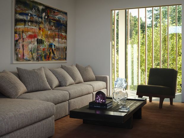 A refined living room with a large neutral-grey sectional sofa, dark rectangular coffee table, and sculptural decorative objects. A colourful abstract artwork forms a feature above the sofa, while full-height timber-framed glazing with vertical screening opens to lush greenery outside. The room has a warm brown carpet and a calm, contemporary atmosphere.
