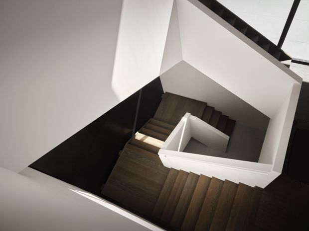 A dramatic overhead view down a modern stairwell, showing multiple flights of dark timber stairs arranged around a central angular landing. White plaster walls and balustrades contrast with black wall panels and dark framing, creating a sculptural geometric composition.