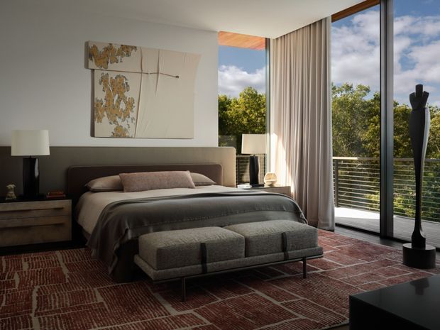 A sophisticated contemporary bedroom with a low upholstered bed, layered neutral bedding, a long tufted bench at the foot, and matching bedside furniture. Floor-to-ceiling glazing opens to a balcony with horizontal metal railings and leafy outdoor views, softened by full-height curtains. A large abstract artwork, sculptural black floor lamp, dark side table, and patterned reddish-brown rug add refined contrast.