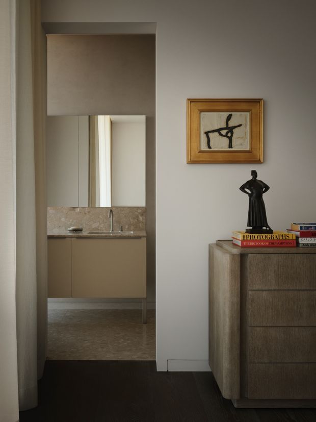 A refined interior view looking through a wide opening toward a compact bathroom or vanity area. The foreground features a warm wood-toned chest of drawers topped with books and a dark sculptural figure, with framed abstract artwork above. Beyond, a floating vanity with a pale stone backsplash and integrated sink sits beneath a large mirror. The space uses muted taupe walls, dark timber flooring, warm timber joinery and soft, understated lighting.