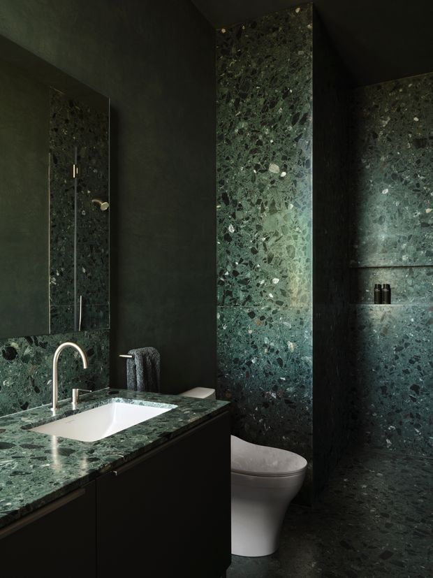 A dark, luxurious bathroom featuring deep green terrazzo or stone surfaces across the vanity benchtop, shower/toilet wall, and floor. A white integrated basin sits in a dark built-in vanity with a curved chrome tap. The image also shows a wall-mounted mirror, a concealed shower area, toilet, recessed shelving, and a charcoal textured wall finish.