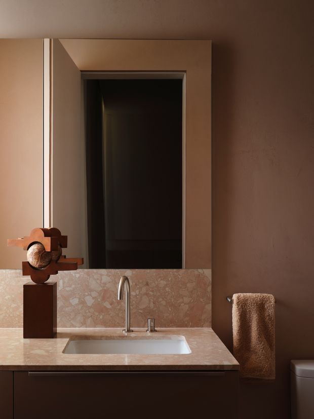 A moody, refined bathroom vanity with a rose-toned natural stone benchtop and matching low splashback. The composition includes a rectangular undermount basin, brushed metal gooseneck tap, wall mirror, dark brown cabinetry, warm brown walls, a sculptural object on the counter, and a textured brown hand towel. A partially visible toilet confirms the bathroom setting.