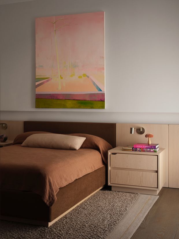 A warm, contemporary bedroom featuring a low upholstered bed with a brown duvet and headboard, pale timber built-in bedside cabinetry, a matching three-drawer nightstand, and a textured neutral rug over dark timber flooring. A large pastel pink and yellow artwork hangs above the bed, with soft wall lighting and understated decor creating a calm, refined atmosphere.