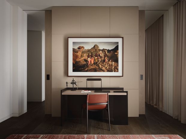 A refined, compact home office centered on a built-in dark desk with a slim task lamp and a red upholstered chair. The room features a large framed artwork on a warm beige paneled feature wall, dark timber flooring, full-height neutral curtains, and concealed or integrated storage framing the workspace. The styling is restrained and contemporary with warm, subdued lighting.