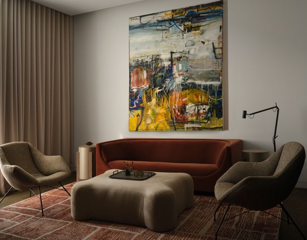 A refined living room with a deep burgundy curved sofa, two sculptural upholstered lounge chairs, and a large rounded ottoman arranged around a low tray. Floor-to-ceiling taupe curtains, a textured geometric rug, a slim black floor lamp, and a vibrant abstract artwork create a warm, sophisticated mid-century-inspired setting.