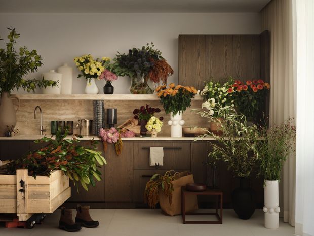 A warmly lit contemporary kitchen or floral studio display with dark timber cabinetry, a pale stone benchtop and matching splashback, and a stainless steel undermount sink with brushed metal tapware. The room is densely arranged with abundant fresh flower arrangements and potted greenery across the benchtop and floor. A timber display crate, small stool, and boots add a rustic working-studio feel, while full-height curtains frame the right side.