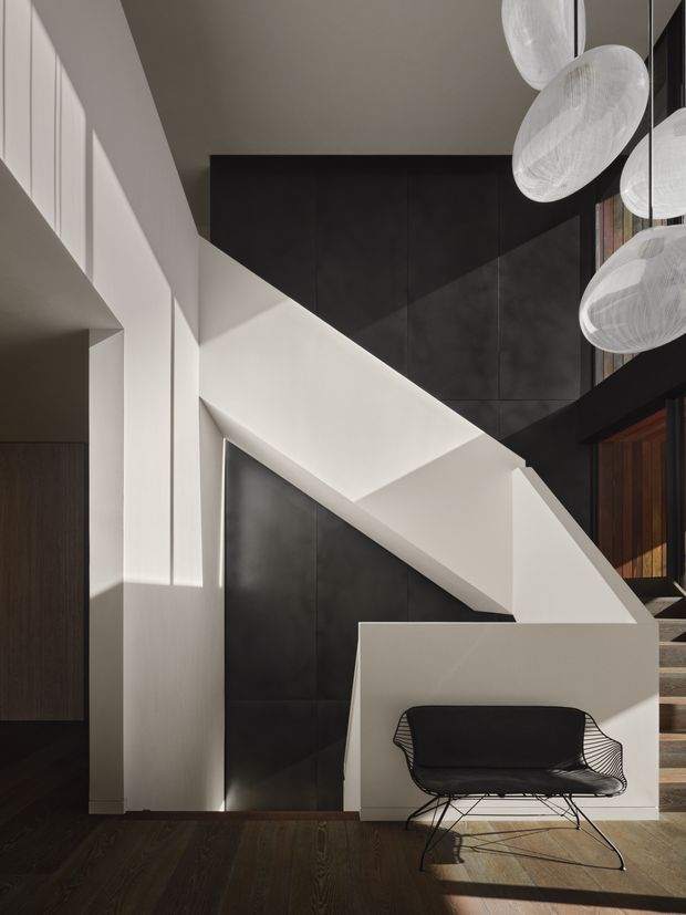 Interior view of a dramatic modern stairway with a crisp white sculptural stair and balustrade set against dark wall panelling. A black wire lounge chair sits in the foreground on dark timber flooring, while clustered translucent pendant lights hang above. Strong sunlight creates geometric shadows across the minimalist space.