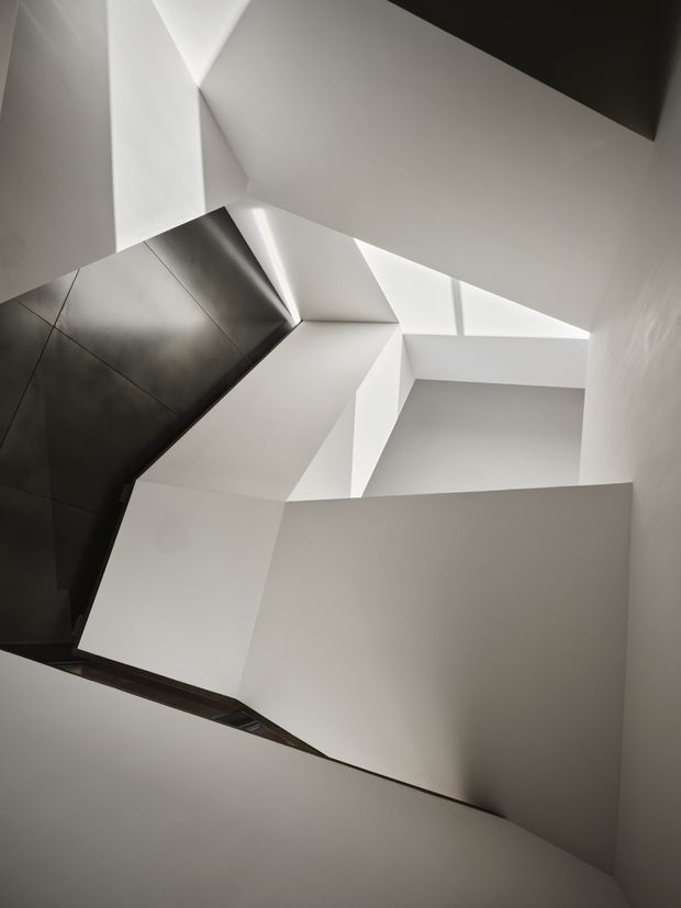 Abstract upward-looking view within a stark, sculptural interior circulation space. Angular white plastered planes and intersecting stair or landing forms create a faceted composition, with a dark reflective wall or cladding panel at left and strong natural light entering through a narrow skylight or clerestory opening. The image is minimal, monochromatic, and architectural, with no furnishings visible.