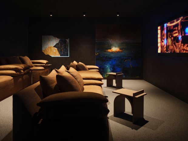 A dark, atmospheric media room or home cinema with rows of deep cushioned lounge seating facing a wall-mounted illuminated screen. Black walls and ceiling, recessed spotlights, textured dark flooring, and several artworks create a moody, luxurious viewing environment. Small sculptural side tables sit between the seating rows.