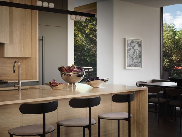 A refined contemporary kitchen and dining area with a large pale stone island, integrated timber cabinetry, black bar stools, a stainless-steel tap, and a dark-framed floor-to-ceiling window. A dining table and chairs are visible beyond the island, with greenery outside.