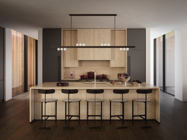 A contemporary kitchen centered on a large timber island with a pale stone or engineered-stone benchtop and five black bar stools. The kitchen features extensive light timber cabinetry, dark charcoal wall cabinetry, integrated appliances, a timber-toned splashback, dark timber flooring, and a long linear pendant light with exposed globe bulbs. Full-height glazing or openings on both sides bring in natural light and reveal timber screening elements.