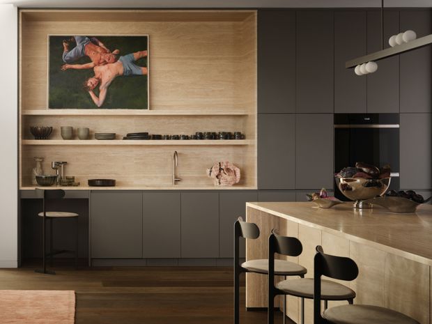 A sophisticated contemporary kitchen with dark matte cabinetry, a large light timber island, integrated appliances, open timber display shelving, and minimalist pendant lighting. Black stools with light upholstered seats line the island, while warm timber flooring and artwork add contrast.