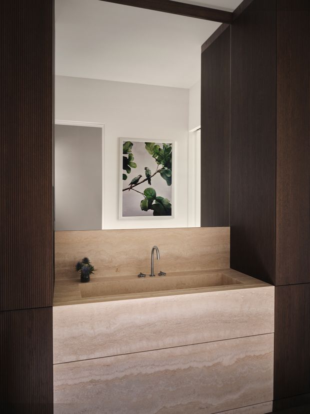 A refined bathroom vanity with a wide integrated stone basin and matching stone backsplash, framed by dark vertically fluted built-in cabinetry. A slim arched metal tap sits above the basin, with a wall mirror reflecting a light neutral interior and framed botanical artwork. The scheme combines warm beige stone, dark timber-toned joinery, and soft white walls.