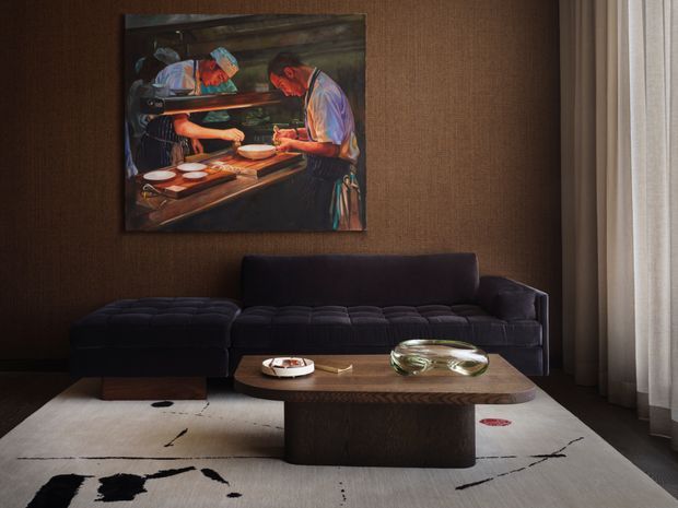 A moody, refined living room with a deep charcoal modular sofa against a textured dark brown wall. A large colourful artwork hangs above the sofa, while a low sculptural timber coffee table sits on a cream rug with bold black abstract markings. Sheer floor-to-ceiling curtains soften the daylight at right, and a glass decorative object and small tray rest on the table.