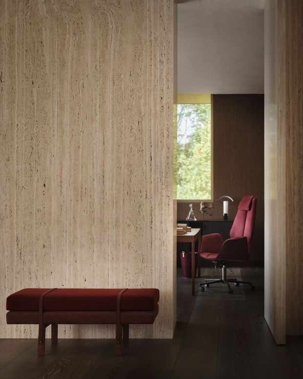 A refined interior view looking toward a compact home office through a textured beige stone partition. The foreground features a deep burgundy upholstered bench with leather straps and dark timber legs. In the office beyond, a matching burgundy swivel chair sits at a desk beside a window, with dark timber flooring, dark wood wall panelling, a slim floor lamp, and a small decorative vessel. The space has a warm, moody, minimalist character with strong material contrast and carefully framed sightlines.
