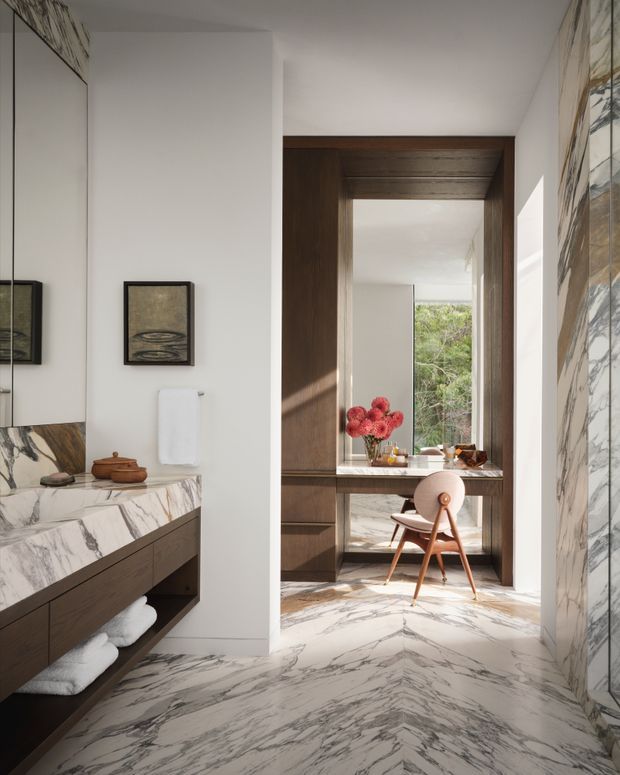 A refined marble-lined bathroom with a floating dark timber vanity, integrated basin, wall mirror, framed artwork, and folded towels. A wide timber-framed opening leads directly to a compact breakfast or dining nook with a marble-topped counter, timber cabinetry, a small round table, and a sculptural wooden chair. Strong natural light enters through the rear window, with greenery visible outside. The space uses dramatic veined marble, warm timber, white walls, and carefully composed contemporary detailing.