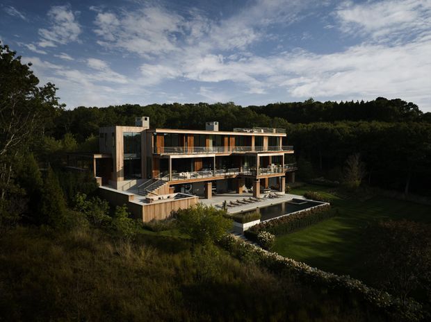 A large contemporary multi-level residence set within a wooded, landscaped site. The building features expansive horizontal glazing, multiple balcony terraces, warm timber-toned exterior elements, flat roof sections, and a stepped design following the terrain. A swimming pool and manicured lawn are visible in the foreground.