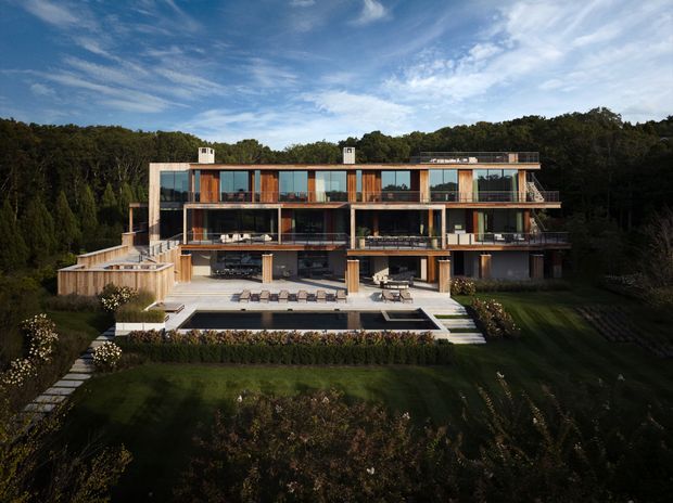 A large contemporary multi-level residence set within a wooded landscape, viewed from the rear across landscaped gardens. The house features extensive floor-to-ceiling glazing, warm timber cladding and trim, dark structural elements, broad terraces and balconies, and a large rectangular swimming pool with surrounding outdoor lounging areas. The composition emphasizes the building’s horizontal form, stepped levels, and strong indoor-outdoor connection.
