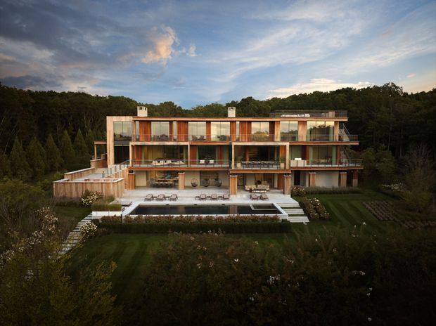 A wide elevated exterior view of a large multi-storey contemporary residence set within a heavily landscaped, wooded site. The building has a long rectilinear form with extensive floor-to-ceiling glazing, warm timber-toned facade sections, balconies, recessed terraces, and a flat roofline. A large swimming pool, paved terrace, outdoor seating, lawns, planted borders, and stepped paths occupy the foreground.
