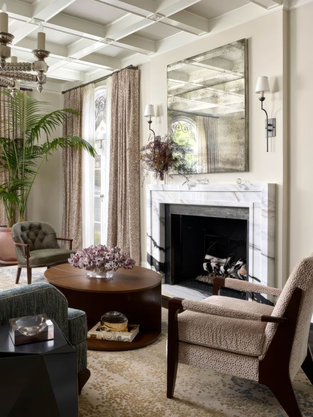 Elegant, traditionally styled living room with a white coffered ceiling, marble fireplace surround, large antiqued mirror, tall curtained windows, and layered seating arranged around a round wood coffee table. The room combines refined neutral finishes with patterned upholstery, a leafy indoor palm, sculptural chandelier, and a soft patterned rug.
