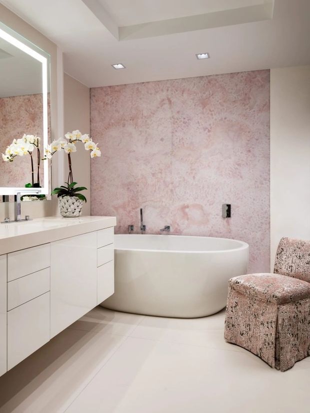 Bright, refined bathroom with a freestanding white oval bathtub, wall-mounted white vanity with integrated basin, large illuminated mirror, recessed ceiling lights, and a pale pink textured feature wall. A plush blush-patterned accent chair and orchid arrangement add soft decorative detail.