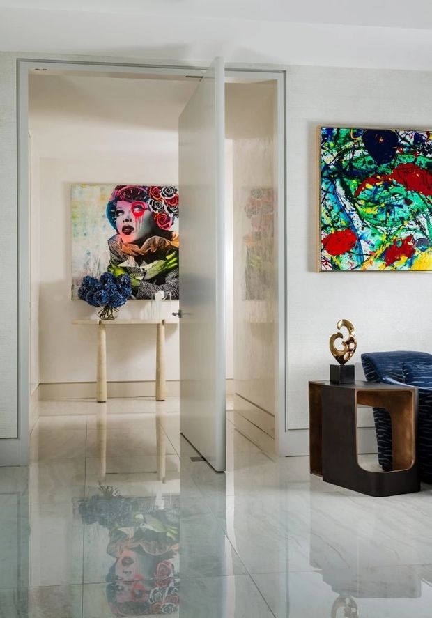 A refined contemporary interior hallway or entry area with highly polished pale stone flooring, white walls, and a glass-panel door opening toward an art-filled room. Colourful contemporary artworks, a sculptural console, decorative object, and dark upholstered seating create a gallery-like, luxe atmosphere. The reflective floor strongly mirrors the artwork and furnishings.