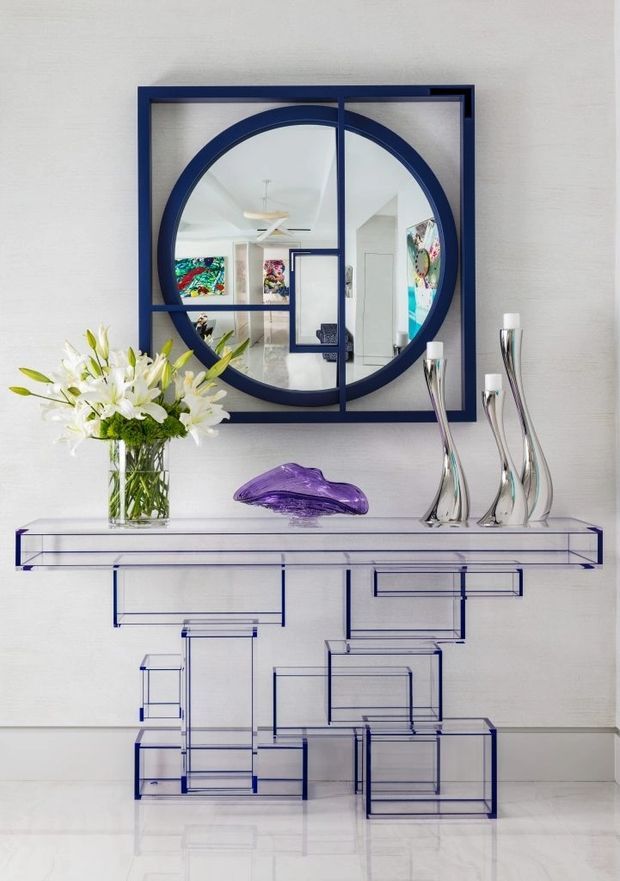 A refined hallway or entryway vignette featuring a transparent glass console table with blue outlined geometric supports, sculptural silver vases, a purple glass ornament, and a clear vase of white lilies. Above it hangs a large navy-framed circular and rectangular mirror, reflecting a bright interior with colorful artwork and contemporary furnishings. The pale textured wall and glossy light floor create a bright, polished setting.