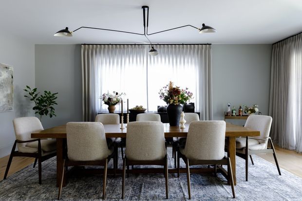 A refined dining room with a long timber dining table surrounded by upholstered dining chairs, large sheer-curtained windows, a contemporary multi-arm pendant light, a sideboard, and floral arrangements. The room has soft grey walls, light timber flooring, and a neutral textured rug, creating a calm, elegant atmosphere.