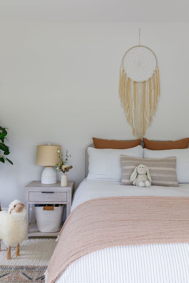 A softly styled bedroom with a white upholstered bed, white-and-grey striped bedding, a blush-pink textured throw, rust-brown cushions, and a striped decorative pillow with a plush rabbit. A pale grey bedside table holds a ceramic lamp and small vase, while a large woven macramé wall hanging decorates the light grey wall. A small white sheep stool and layered rugs add playful, relaxed character.