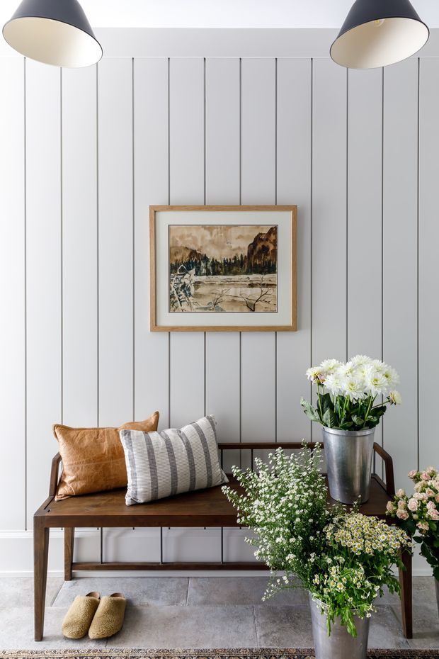 A styled entryway or hallway vignette with a dark timber console bench against a light grey vertical-panelled wall. The bench holds striped and tan cushions, while bouquets of white and pale flowers sit in metal and ceramic vessels. Two large black pendant lights hang overhead, and a framed landscape artwork is centred on the wall. Grey tiled flooring and a partially visible rug complete the calm, traditional-contemporary interior.