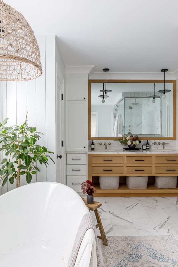 Bright, refined bathroom interior featuring a freestanding white bathtub in the foreground, a double timber vanity with undermount basins, brass-toned fixtures, large framed mirror, marble-look floor and shower surfaces, white cabinetry, and woven pendant lighting. A leafy indoor plant and soft neutral accessories add an organic, relaxed feel.